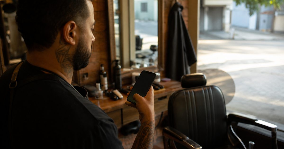 Barbeiro olhando o celular ao lado da cadeira de barbearia, conferindo a agenda online do dia