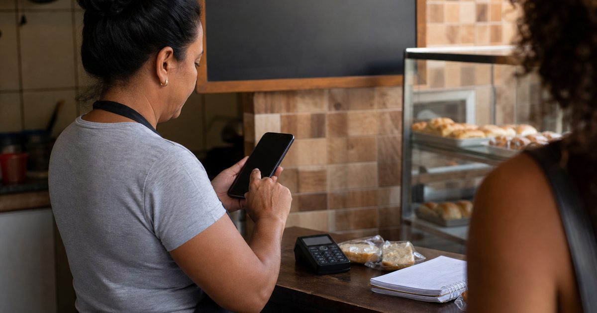 Dona de lanchonete no balcão registrando uma venda no celular, com a loja ao fundo — PDV para pequeno negócio